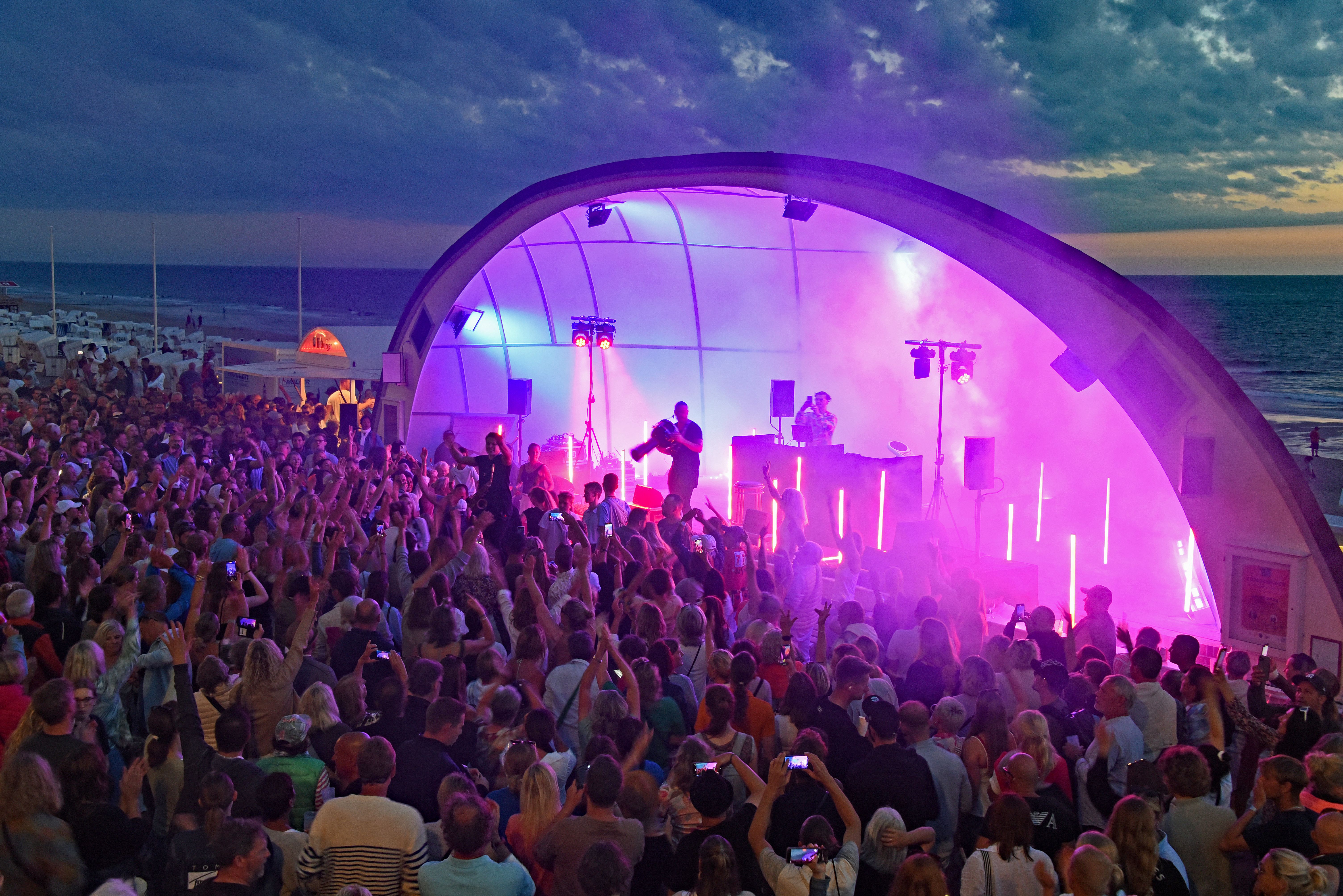 Blick auf die beleuchtete Bühne, auf der eine Band auftritt, viele Menschen feiern vor der Bühne