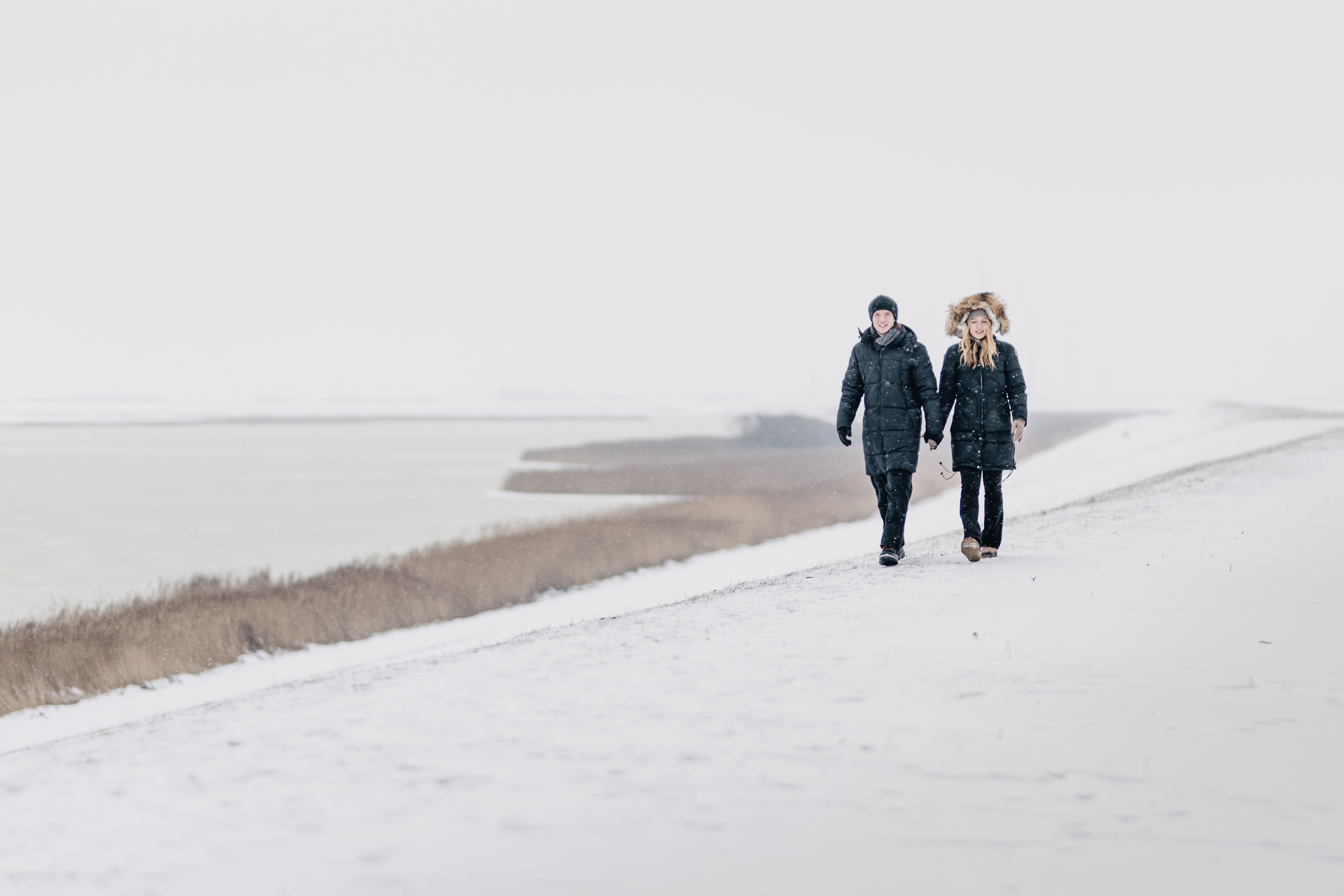 Ein Paar geht im Winter an einem Deich an der Nordsee Schleswig-Holstein spazieren. Es schneit und am Boden liegt bereits etwas Schnee.